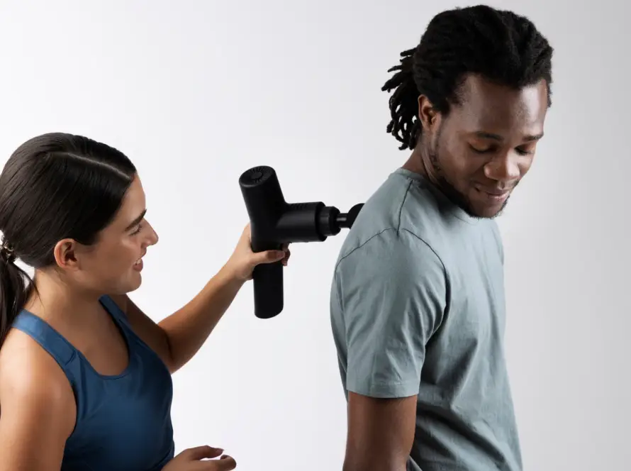 Woman using a massage gun on a man’s upper back while he smiles during home therapy session