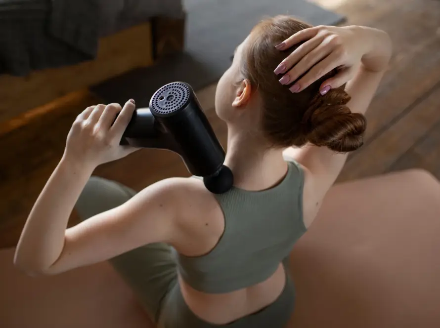 Woman using a massage gun on the back of her neck while seated on a yoga mat