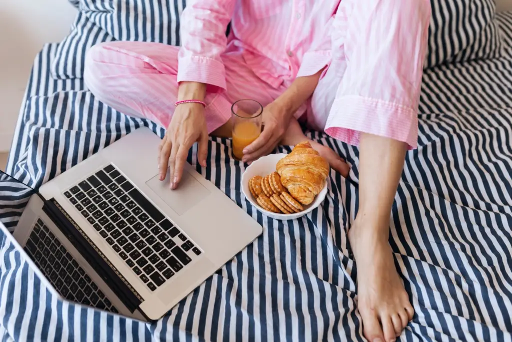 A person enjoying their morning routine by working on a laptop in bed with a breakfast of a croissant and orange juice.